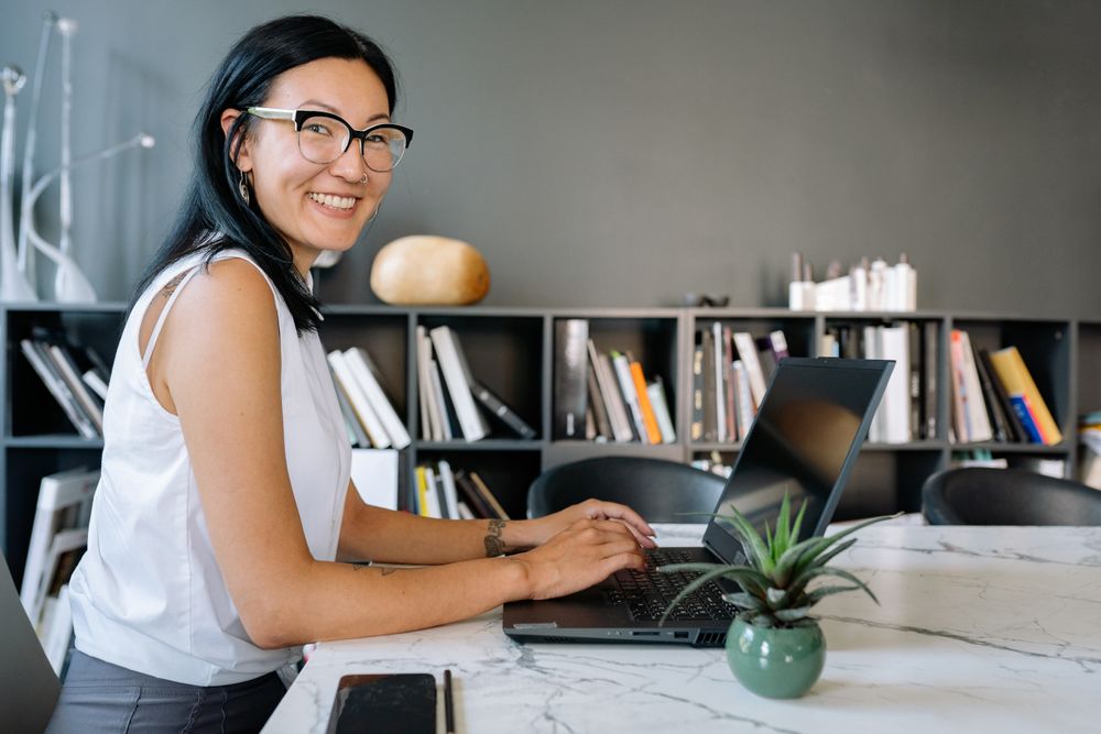 A smiling woman with glasses using a laptop in front of bookshelves