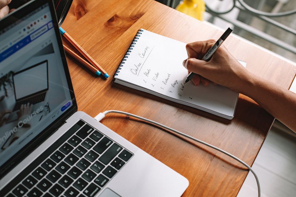 A person writing notes in a notepad beside a laptop