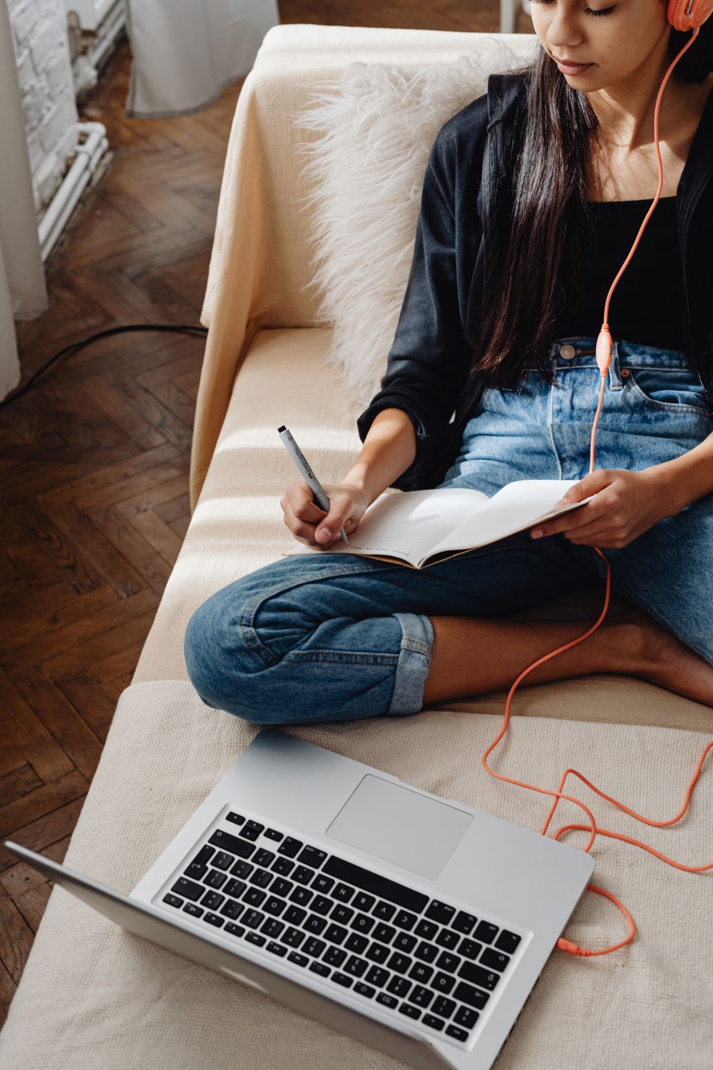 A student on a couch with headphones, a notebook and a laptop